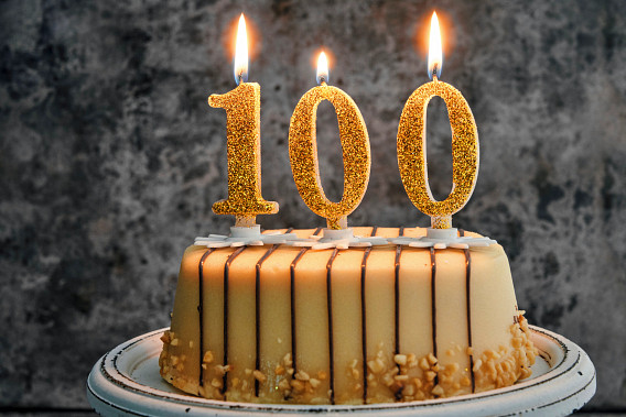 A celebratory cake with three glittery gold candles forming the number "100," all lit and glowing brightly. The cake has a striped design with a smooth beige frosting, chocolate drizzle on the top, and nut garnish around the base. It is placed on a white plate with a rustic background, creating a festive and elegant presentation.