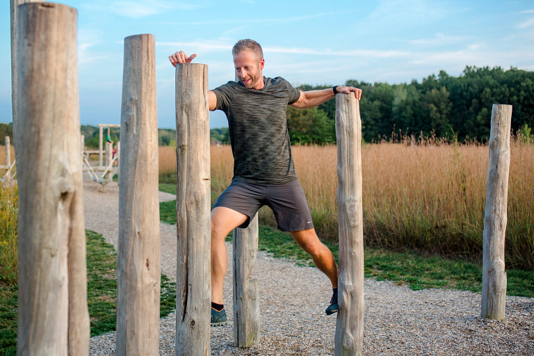 A man exercises outdoors by stepping between tall wooden posts on a gravel path. He grips the posts for balance while moving forward, with grassy fields and trees in the background under a clear sky.
