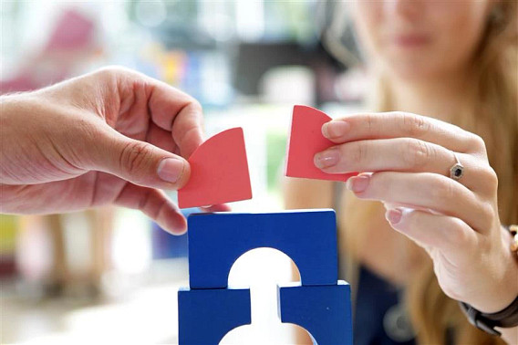 Two hands holding red puzzle pieces above a blue block with cut-out shapes,    appearing to collaborate in fitting the pieces together.