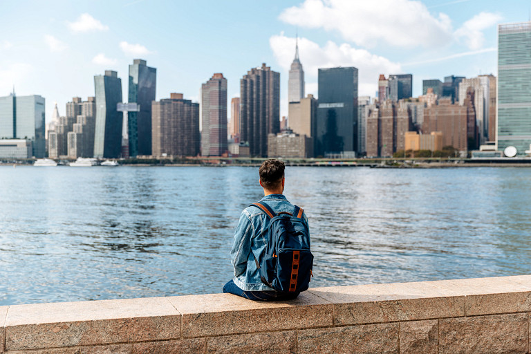 Young man with backpack sitting and looking at New York City skyline on a sunny day, USA