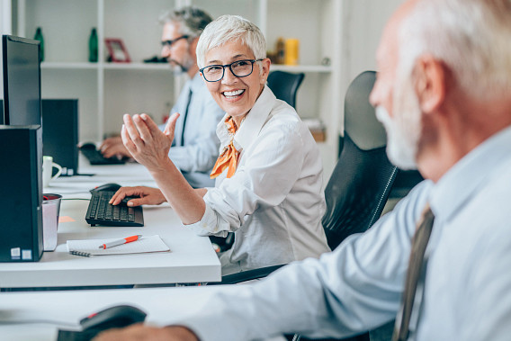 A cheerful office scene with a senior woman sitting at a desk, smiling and engaging in conversation with a male colleague. She is wearing glasses, a white shirt, and an orange scarf. The background shows another man working at a computer in a modern office with shelves and decor. The atmosphere conveys collaboration and professionalism.