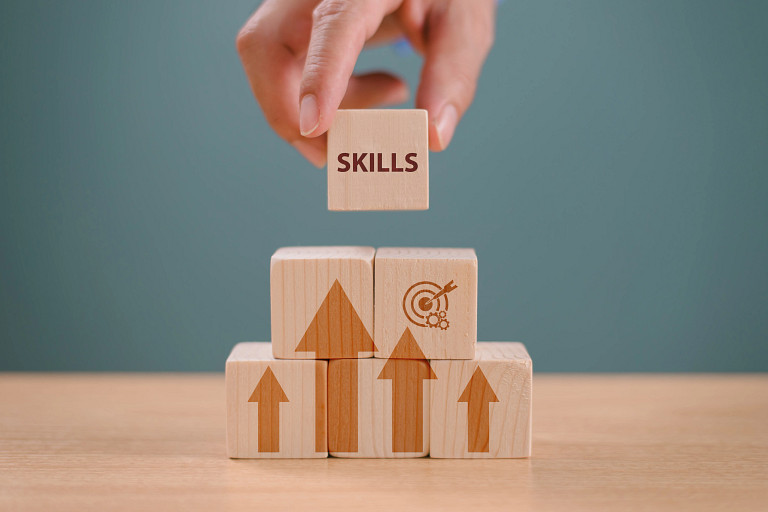 A hand places a wooden block labeled “SKILLS” on top of stacked wooden cubes with upward arrows and a target icon. The blocks sit on a table against a plain background, symbolizing growth, development, and goal-oriented learning.