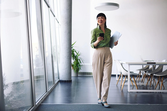 Young Asian businesswoman walking in modern office holding documents and smartphone