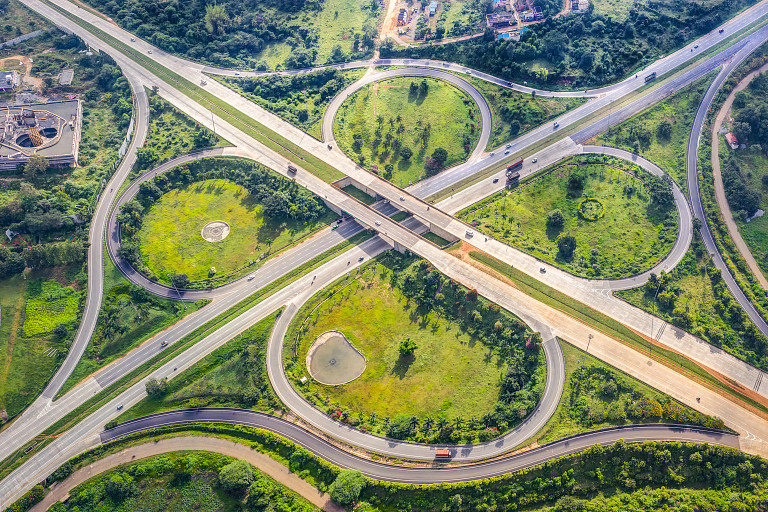 Aerial view of a green, landscaped highway interchange with multiple roads forming loops and crossings in Bangalore, India. The scene features grassy areas, small ponds, and scattered trees, surrounded by forested landscapes and structures in the distance. Vehicles are visible on the roads.