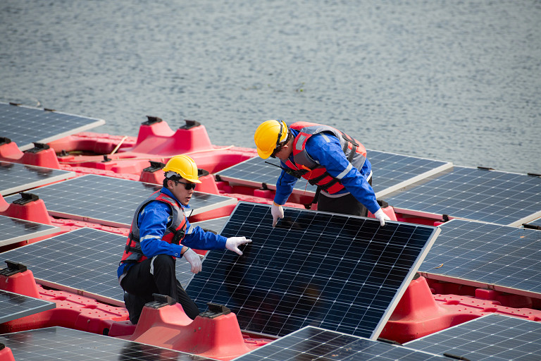 Two workers in yellow helmets and life vests install or inspect solar panels on a    floating platform over water. The panels are mounted on red buoyant structures,    showcasing a floating solar power system.