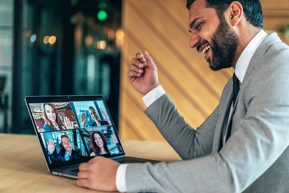 A man in a suit is sitting at a desk, smiling and gesturing during a video call on his laptop. The laptop screen shows four participants in a virtual meeting, each waving or smiling. The background features a modern office setting with soft lighting and wooden paneling. The scene conveys a professional and collaborative environment.