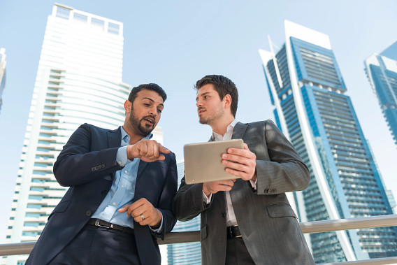 Two businessmen in formal attire are standing outdoors in a modern cityscape with tall skyscrapers in the background. One man is gesturing while the other holds a tablet, suggesting a discussion or collaboration on work. The scene conveys professionalism and teamwork in a corporate setting.