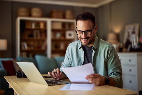man sitting in home office smiling at documents next to laptop
