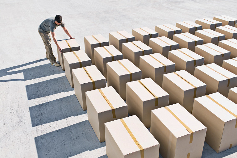 A person arranges sealed cardboard boxes laid out in neat rows on a large concrete surface, viewed from above. The boxes are evenly spaced, creating a grid pattern with long shadows cast in bright sunlight.
